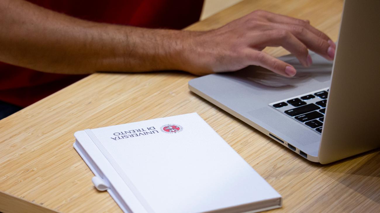 particolare mani con laptop e agenda con logo Università di Trento